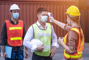 Woman in hardhat doing temp checks on two men with hardhats and COVID masks